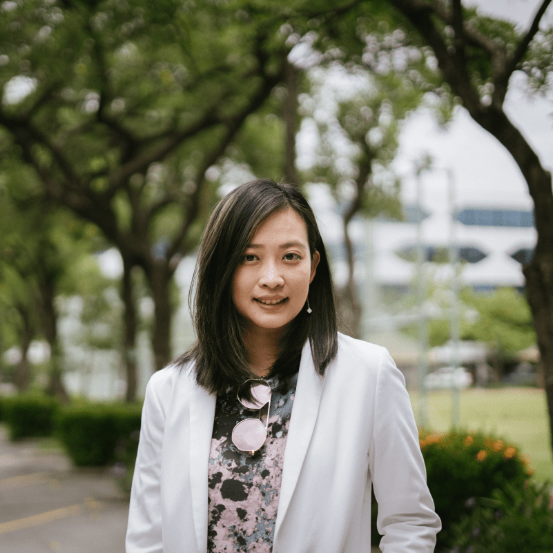 Thitirat is standing outside in a park, wearing a light coloured blazer and a patterned blouse.