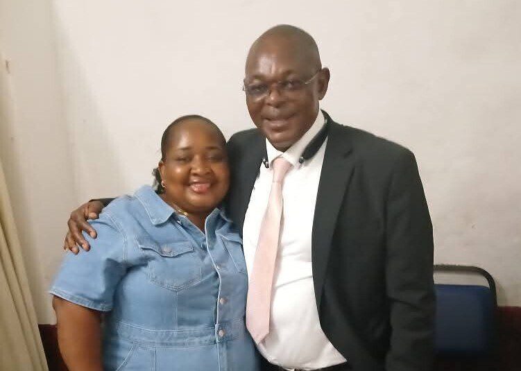 Two smartly dressed people standing side by side with their arms over each others shoulders and smiling.