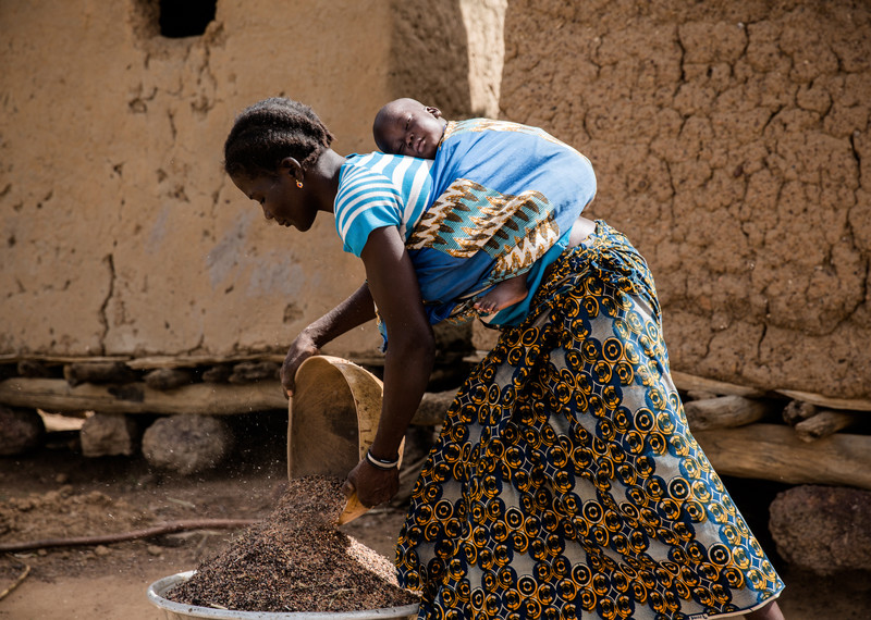امرأة تحمل طفلها على ظهرها وتقوم بتفريغ الحبوب كجزء من الحياة اليومية في قرية لابيين، بوركينا فاسو، يونيو/حزيران 2018 © Sophie Garcia | hanslucas.comSophie Garcia www.sophiegarcia.net