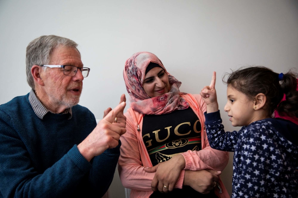 a man with white hair, a woman wearing a pink headscarf and a young girl wearing a hoodie with stars. The man and the young girl look like they are discussing something.