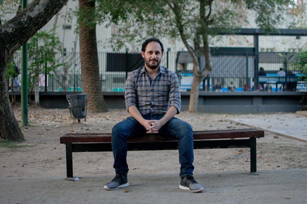 A young man sits on a park bench with his hands across his lap. He is centered in the frame.