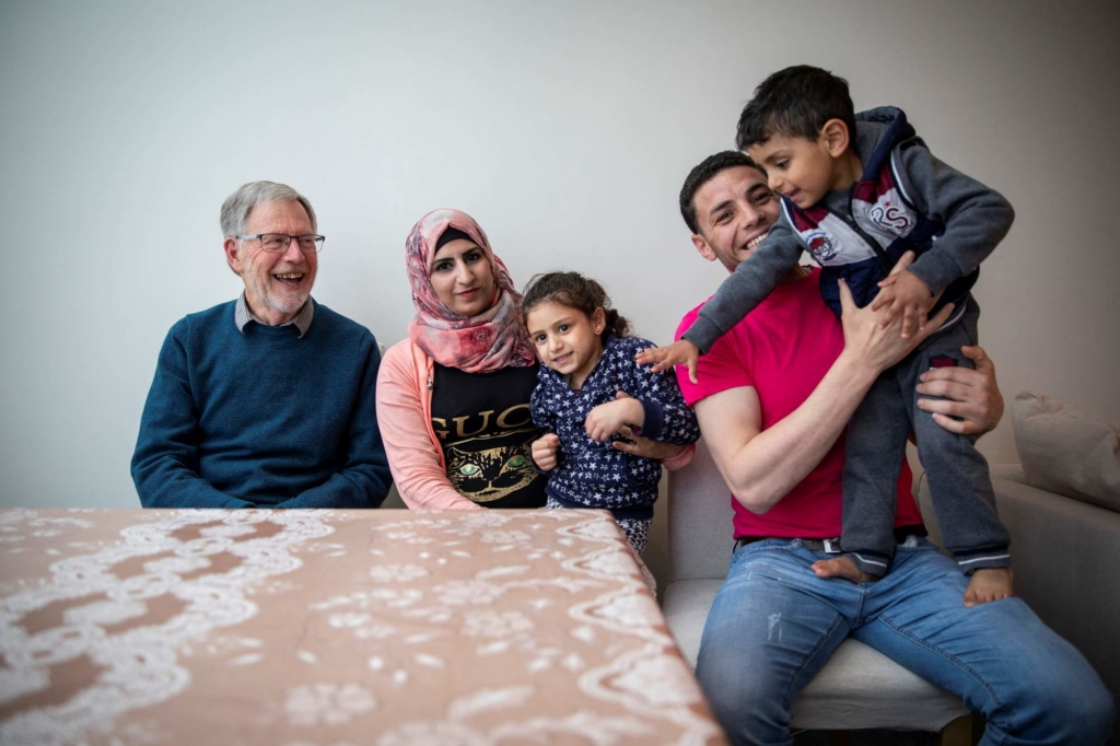 John sits with Rahaf, Monther and their children for a portrait. The young son is playfully standing on his father's lap and is reaching down for something on the table in front of them. 