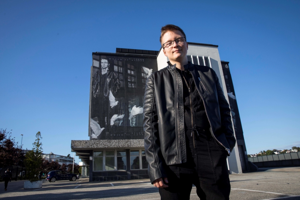 Sakris stands for a portrait in front of a large mural that he is featured in. He is wearing a black leather jacket, a black shirt and glasses. 