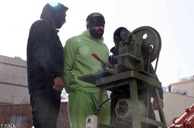 A blindfolded man wearing green standing between two people dressed in black wearing balaclavas. The man is facing an amputation machine.