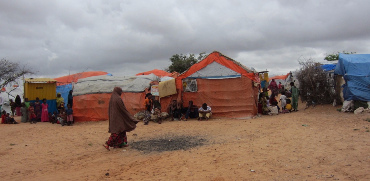 In the foreground, a woman in Muslim outfit walking towards some makeshift shelters. In the background, women and children seated outside the makeshift shelters.