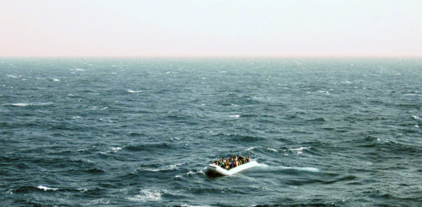 The top third of the screen shows a white sky. The bottom two thirds shows the blue sea. In the bottom right of the image, we can see a small rubber vessel with refugees in it.
