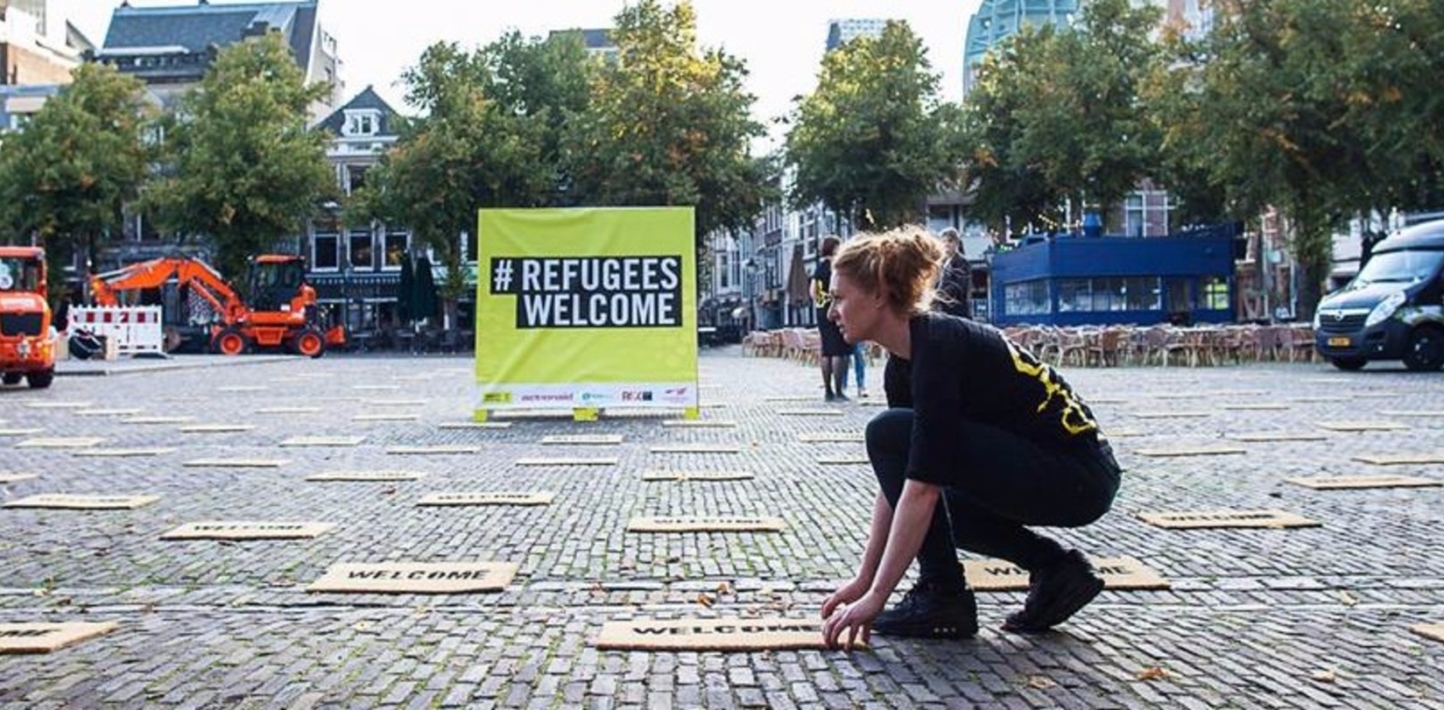 A large 'refugees welcome sign is visible in the background of a large cobbled square. In the forground an amnesty activist places signs saying refugees welcome on the ground.