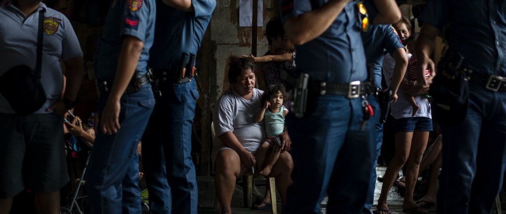 A woman holds her child as she faces a line of police officers. There is a look of despair and exhaustion on her face. 