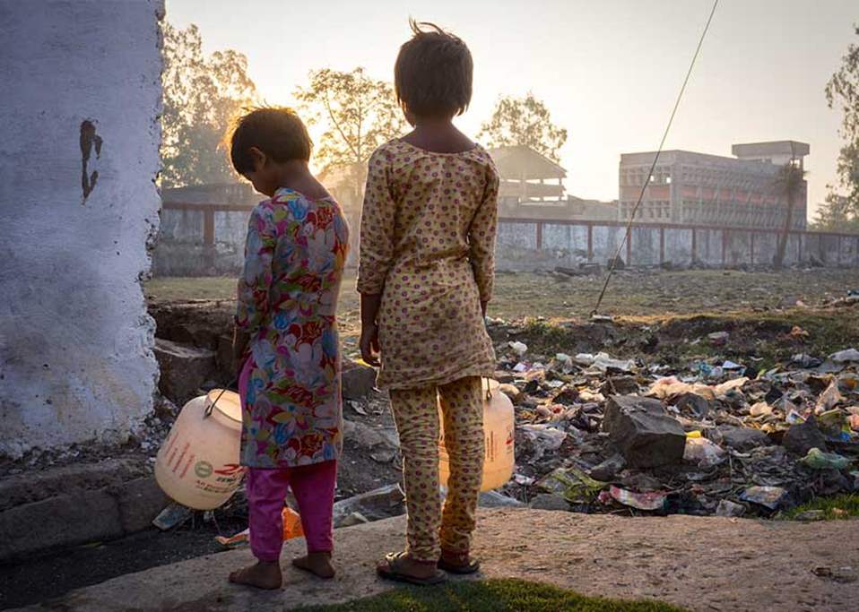 two children stand facing a factory and carry white buckets. 