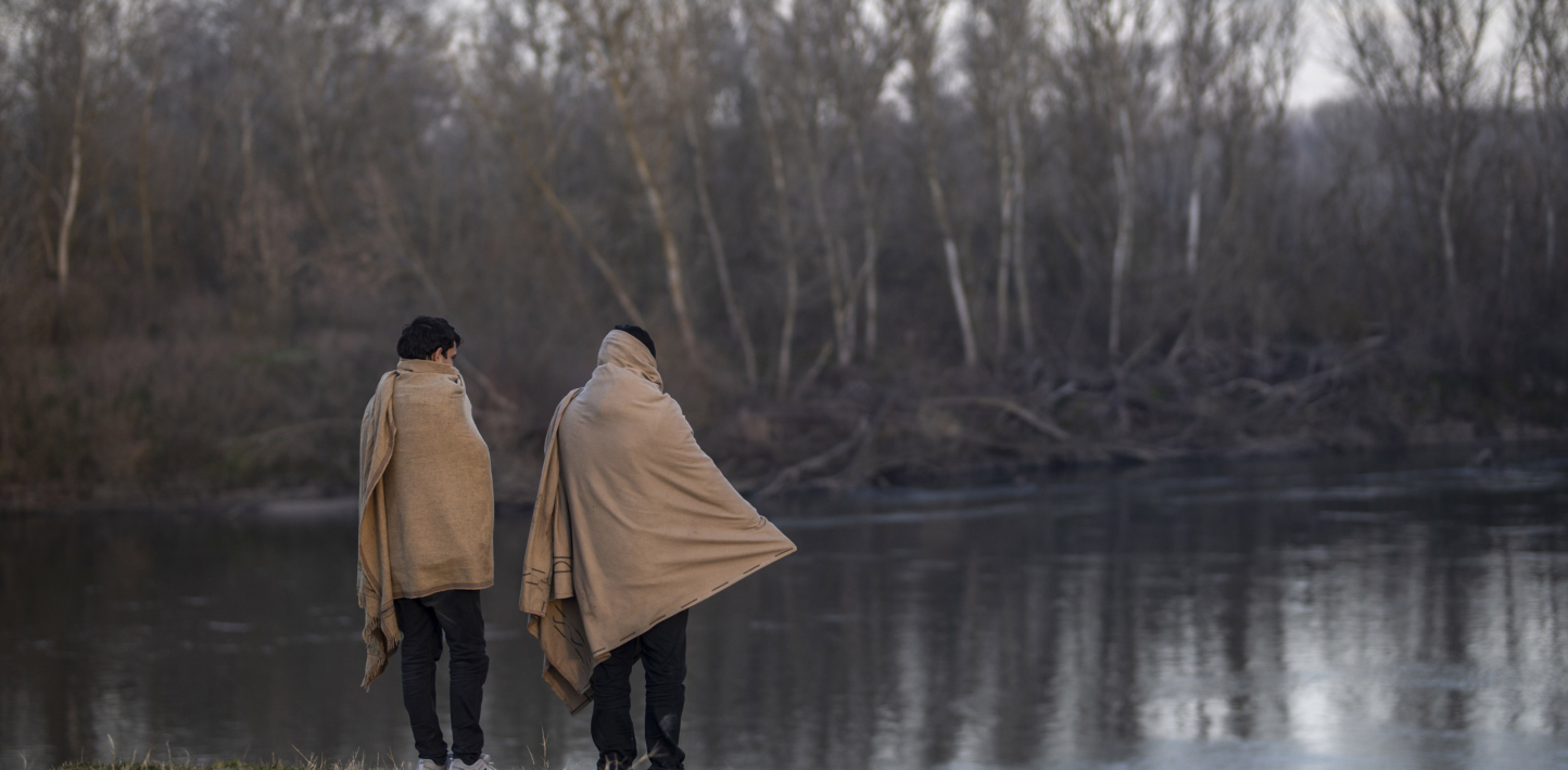 Two people with large blankets over their shoulders standing by a lake in a forest.