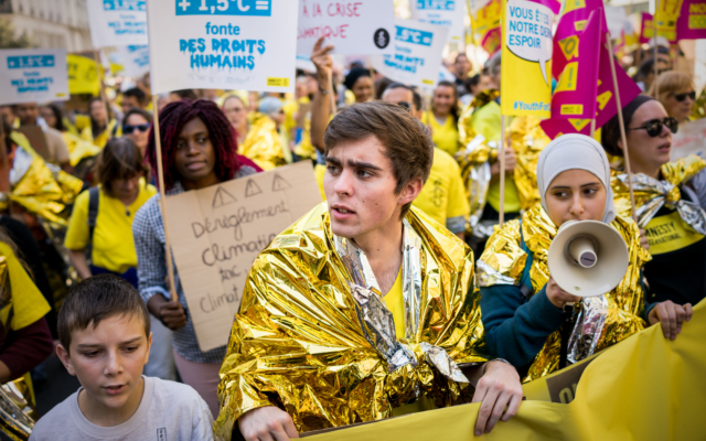 a group of young people attend a protest. Some of them are draped in a gold shiny fabric. They are holding megaphones and protest signs 