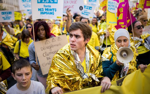 a group of young people attend a protest. Some of them are draped in a gold shiny fabric. They are holding megaphones and protest signs