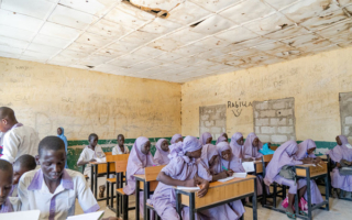 children sit in a classroom, working in groups on an assignment. They are wearing a purple and white uniform.