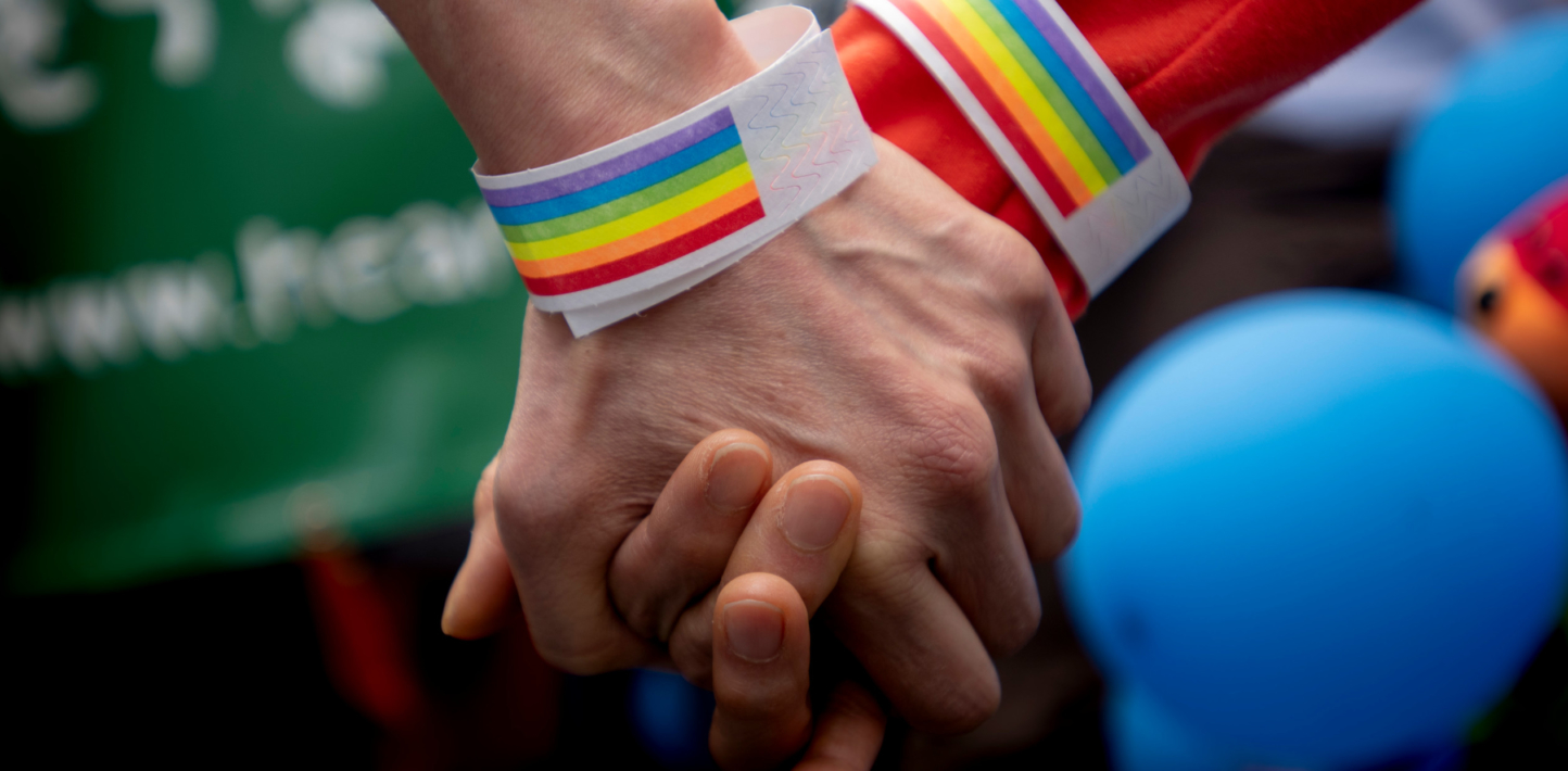 Two hands holding each other with rainbow wristbands.