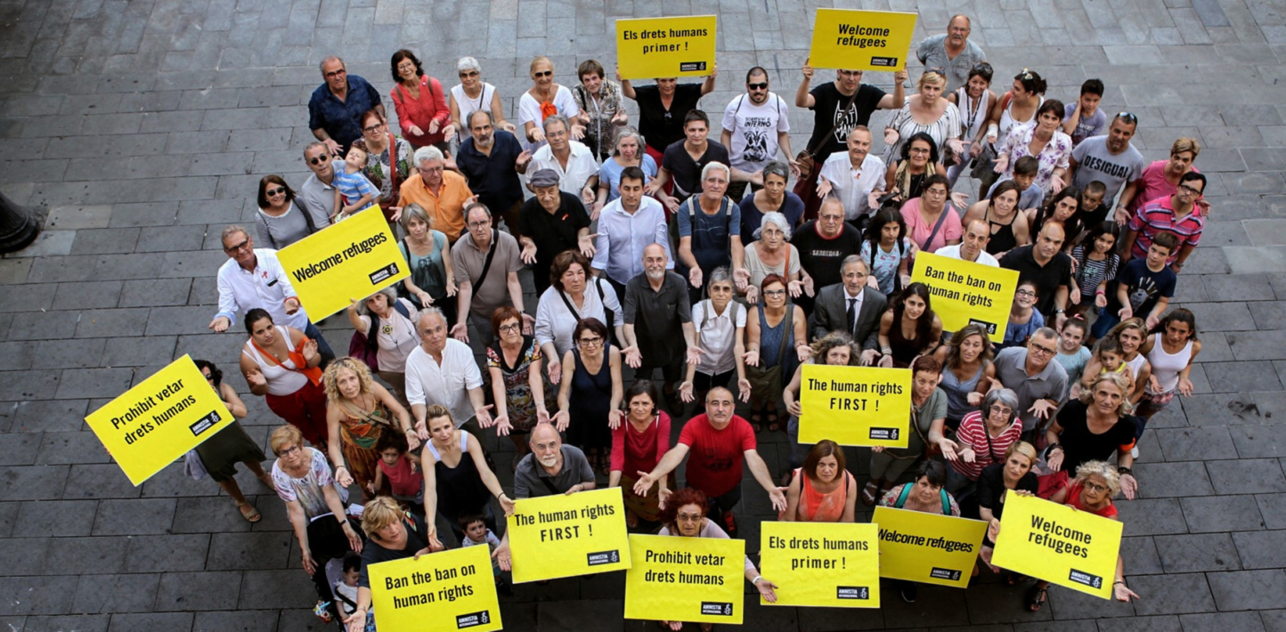 a birds eye view photo of a group of amnesty activists. Some of them have their arms open wide and others are holding signs that read slogans including Welcome Refugees or Ban the Scan.