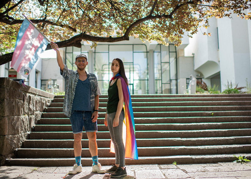 Melike and Özgür stand for a portrait. Melike is wearing a rainbow flag over their shoulders and Özgür is waving a trans pride flag. 