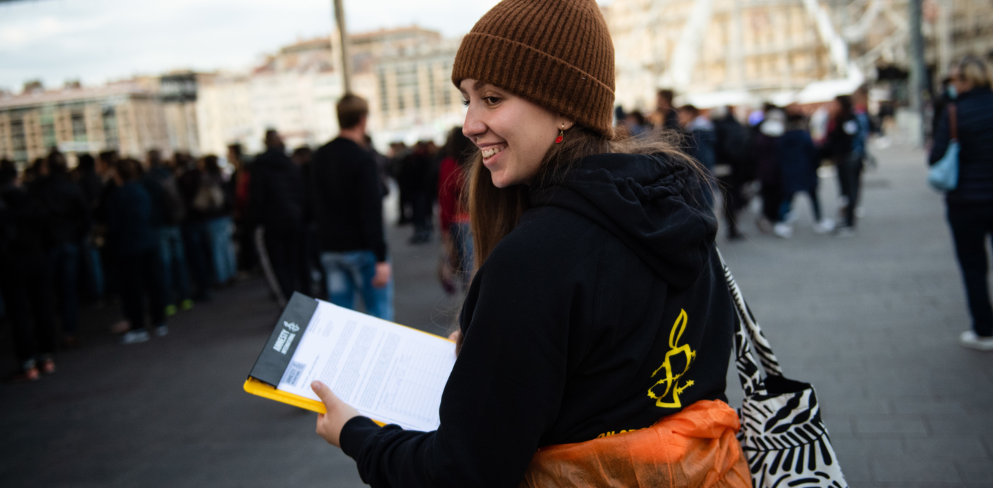 A person turning backwards and smiling, holding a clipboard, with the Amnesty International logo on their hoodie.