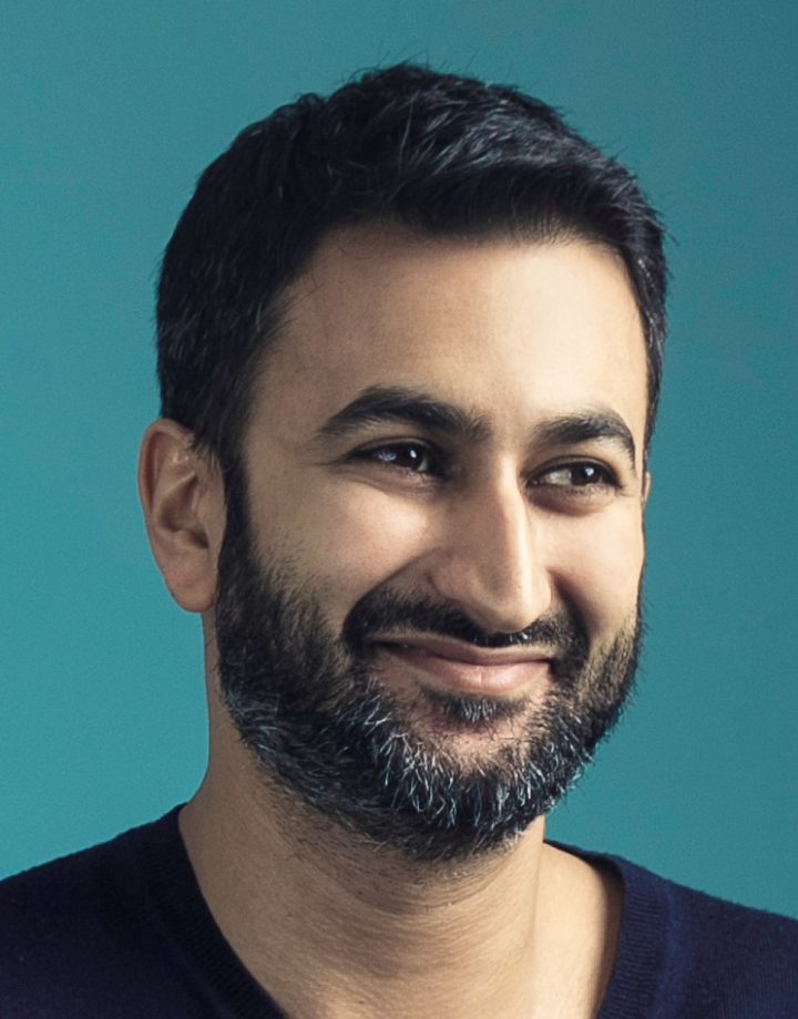 Mandeep Singh, in front of a teal background. He is wearing a black shirt. 