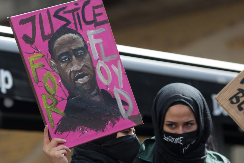 A woman in a facemask and hook holds up a painting of George Floyd that reads "Justice for Floyd"