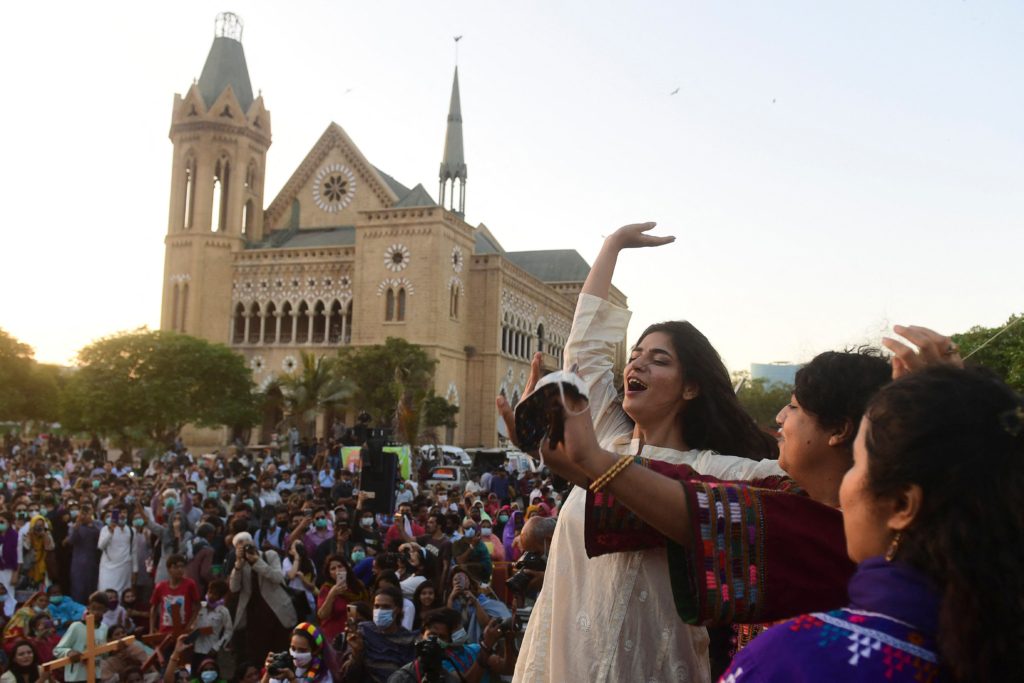 three activists are at the forefront of an image of a large protest in Karachi. They appear to be celebrating with their arms waving joyously. 