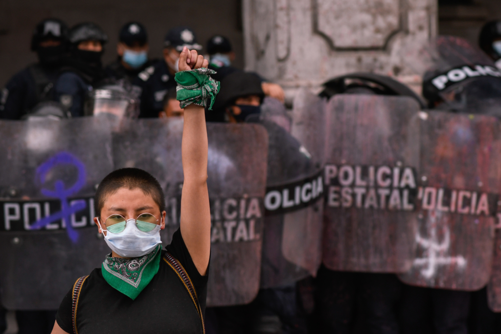 A person wearing a white face mask, green tinted sunglasses and green bandanas around their neck and wrist stands defiantely infront of a line of police with riot shields. They are participating in a protest in Toluca, Mexico, demonstrating against gender-based violence, femicide. 