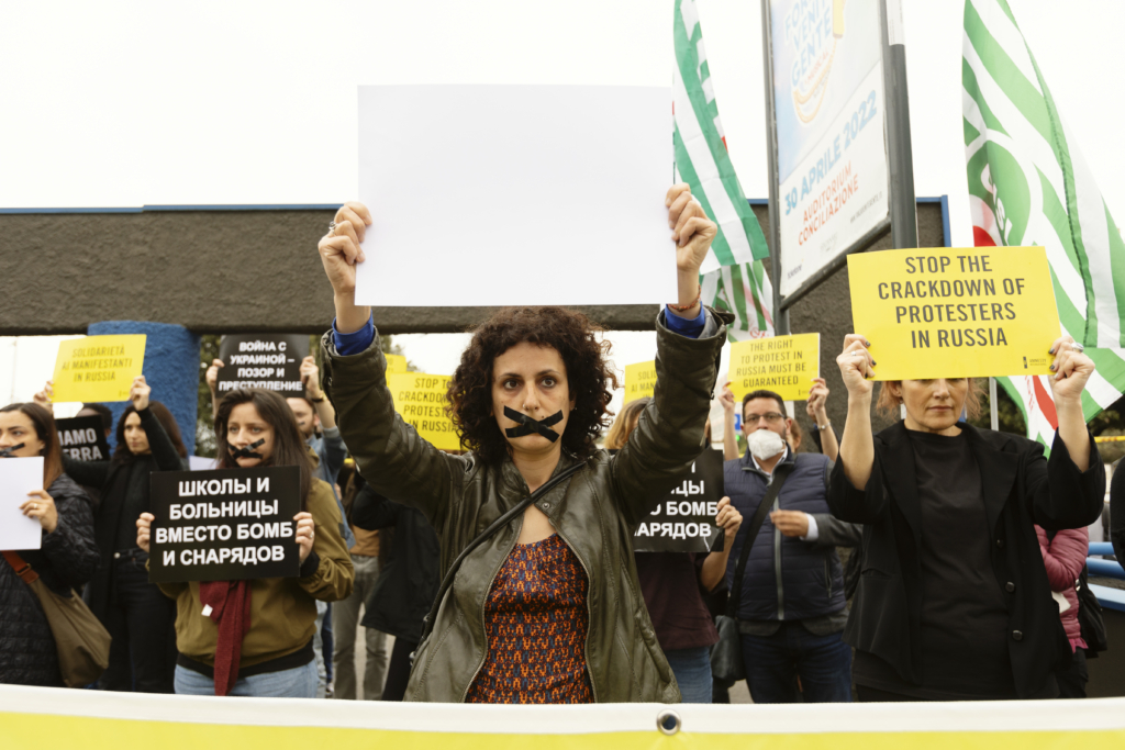 Des militant·e·s d’Amnesty International tiennent des pancartes jaunes et noires, certaines en anglais, d’autres en russe. Plusieurs militantes ont la bouche fermée par du ruban adhésif noir. 
