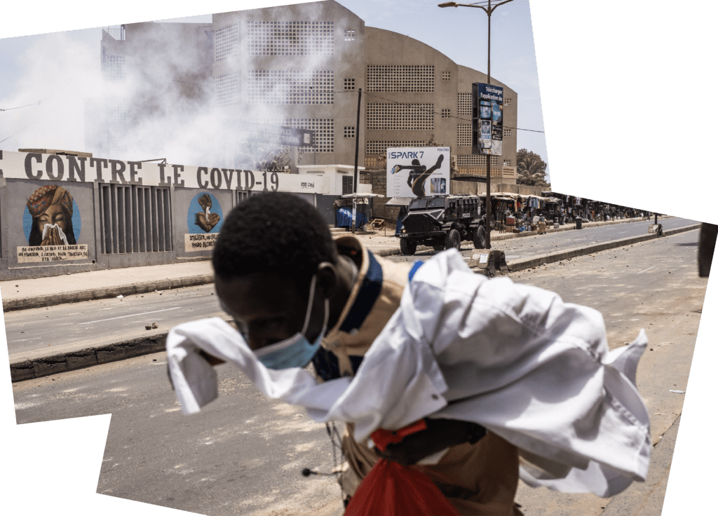  A man runs from rocks and teargas during a protest in Dakar, Senegal. You can see armed vehicles and a light smoke behind him. 