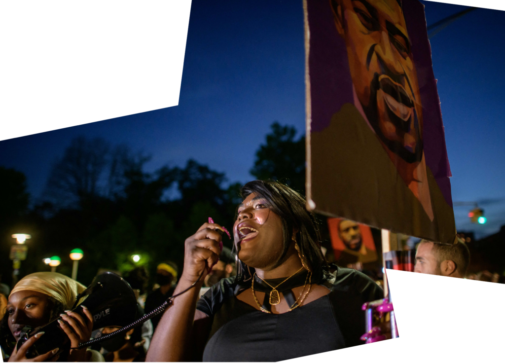 a woman holds the microphone of a megaphone to her mouth as she speaks. She is at a crowded protest and is holding a placard with a painted image of George Floyd 