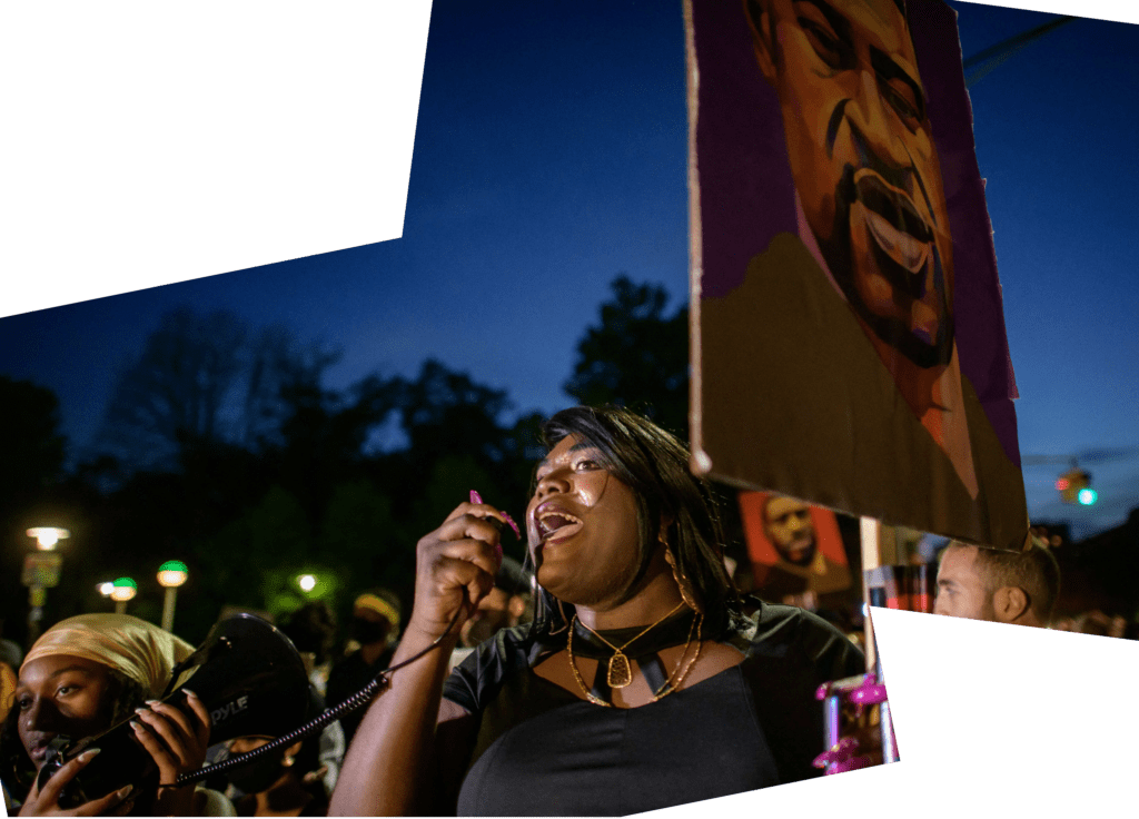 a woman holds the microphone of a megaphone to her mouth as she speaks. She is at a crowded protest and is holding a placard with a painted image of George Floyd 