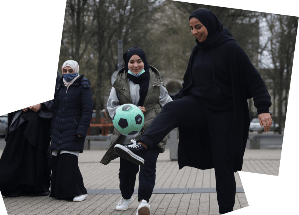 a group of women, all wearing headscarves, kick around a green and black football. They look gleeful as they play. 