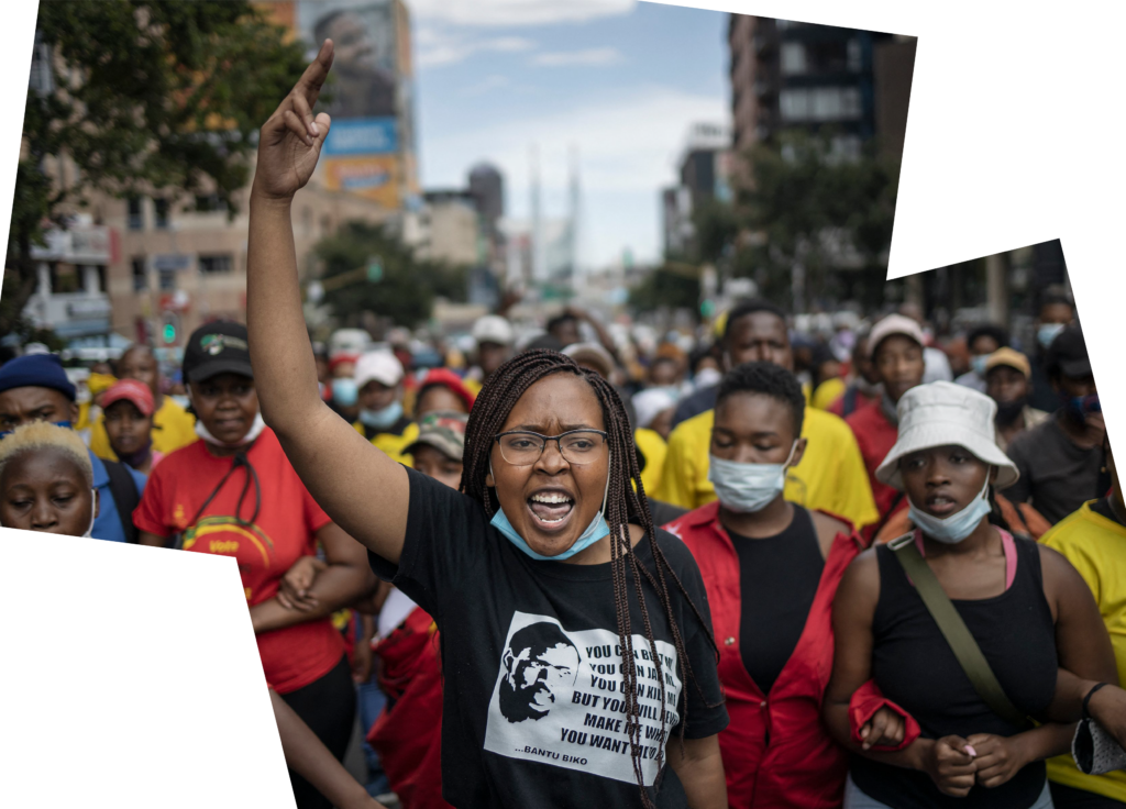 Une femme se tient devant une foule de manifestant·e·s, le doigt levé. Elle porte des lunettes et un T-shirt noir