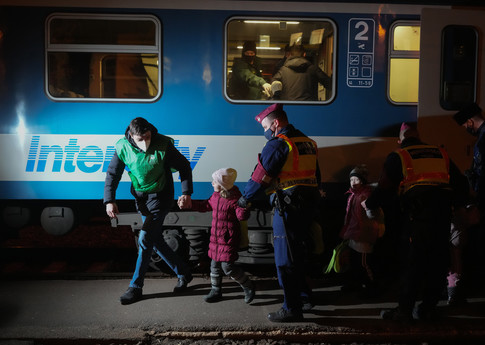 A group of children, all in winter clothing, are greeted by adults in bright vests and face masks as they step off a blue train.