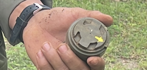 a close up show of a hand holding an M-14 antipersonnel landmine. It is small, about the size of the person's palm and dark green. 