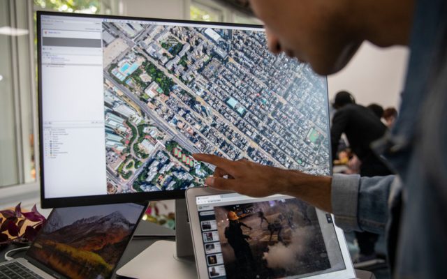 Someone is pointing at a large computer screen that shows maps and satellite imagery. There are several other screens on the desk and you can see others working around them.