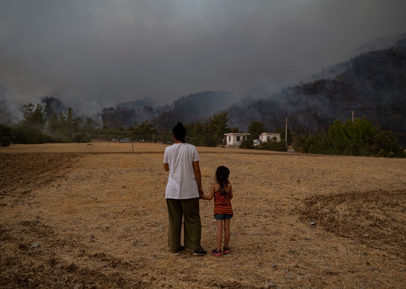 A woman and her daughter stand on a dry sandy road, their faces are turned from the camera as the look at a dark, hazy skyline where wildfires are raging. 