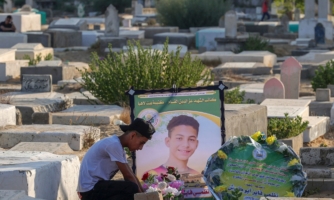 a young man wearing a black baseball cap kneels in front of a grave in a space filled with tombstones. Next to the grave is a framed image of another young man who looks to be of a similar age.