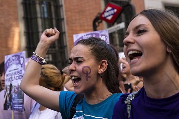 two women are in the focus of a photo taken at a protest. The one on the left has a purple Venus symbol painted on her cheek and has one of her hands in the air in a fist.