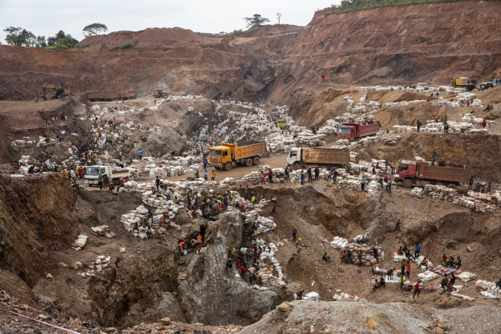 a drone camera image of a cobalt mine in the DRC. The area is surrounded by trucks and hundreds of workers and stacks of bags filled with ore. 
