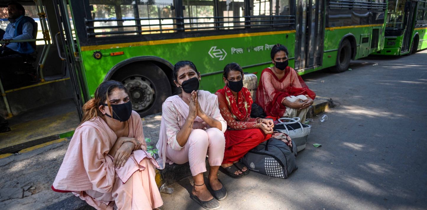 Four transgender people sit together, one looks as though they may be pleading for money. They are all four wearing facemasks.