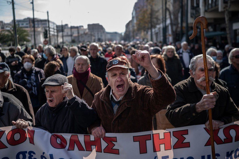 a crowd of older people attend a protest about pension security. Those at the front are holding a sign and you can see them cheering.