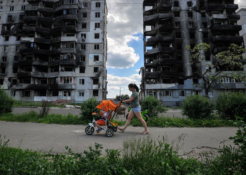 A woman walks past bombed out buildings pushing a child's pushchair.