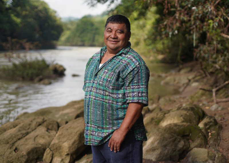 Bernardo Caal Xol standing by a vibrant river in a forest. He is smiling and wearing a blue, purple and green patterned shirt 