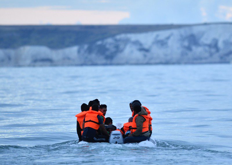 A small overcrowded boat sits low in the water, motoring towards white cliffs in the distance.