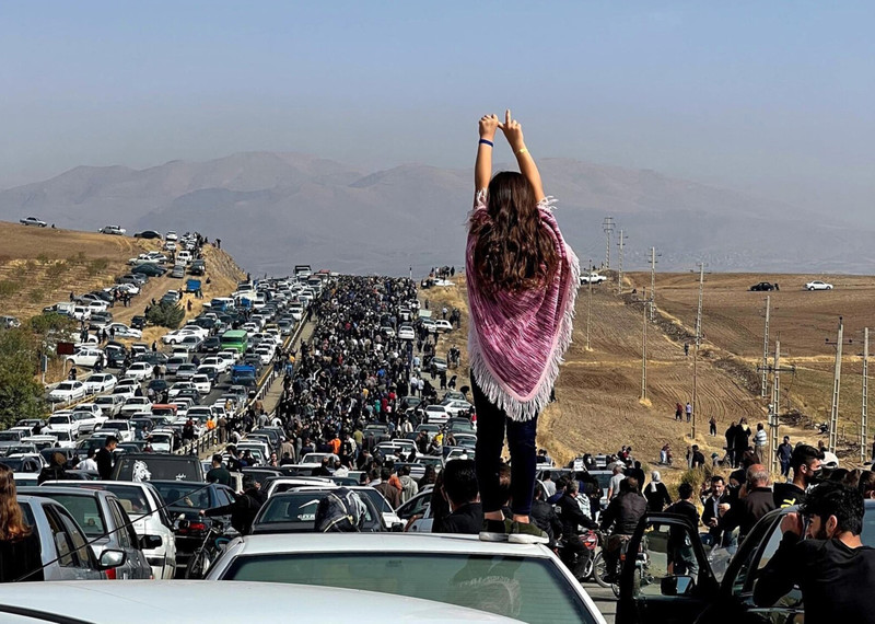 A woman stands defiantly with her hands in the air, a long line of cars and people stretching out into the distance.