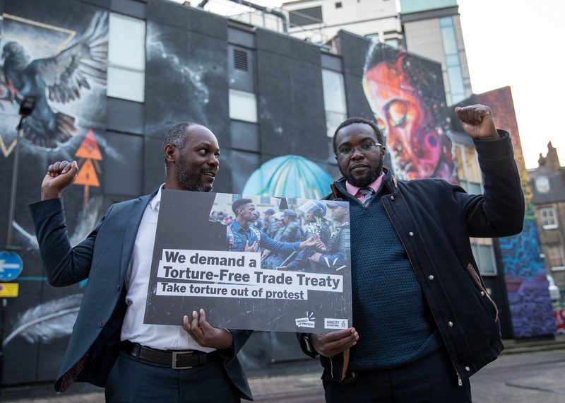 Two men hold a poster with the words 'we demand a torture-free trade treaty', their other hands with fists raised. 