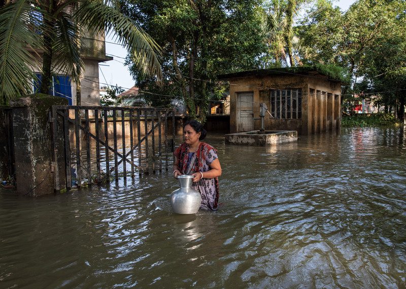 A woman wades through high water holding a large jug.