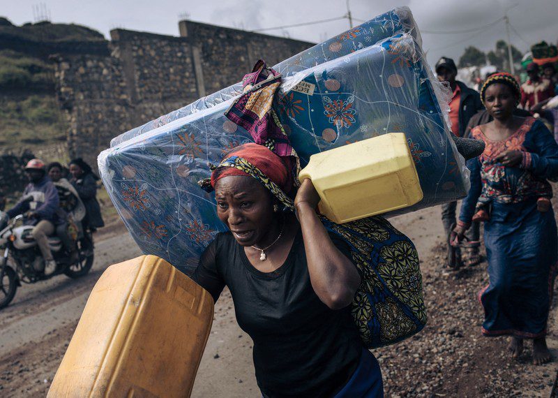 A woman carries luggage on her back and shoulders, looking panic stricken, followed by others.