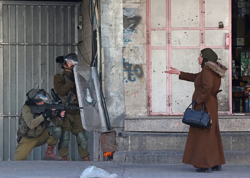 Two people heavily armored, behind shields, point their weapons towards a woman with an outstretched hand.