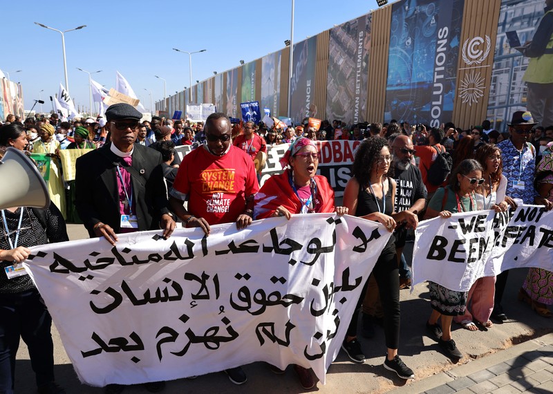 A large crowd of protesters march forwards holding a white banner with black writing. The slogan on the banner reads in Arabic: \" There is no climate justice without human rights. We have not been defeated\"