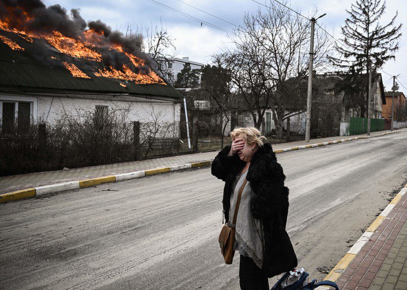 A woman covers her face in anguish, looking on as a house is engulfed by flames.